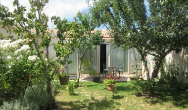 Extérieur de la maisonnette, jardin fleuri et arboré, avec table et chaises sur la terrasse, à l'ombre d'un grand parasol Maisonnette - Sylvette Joubert